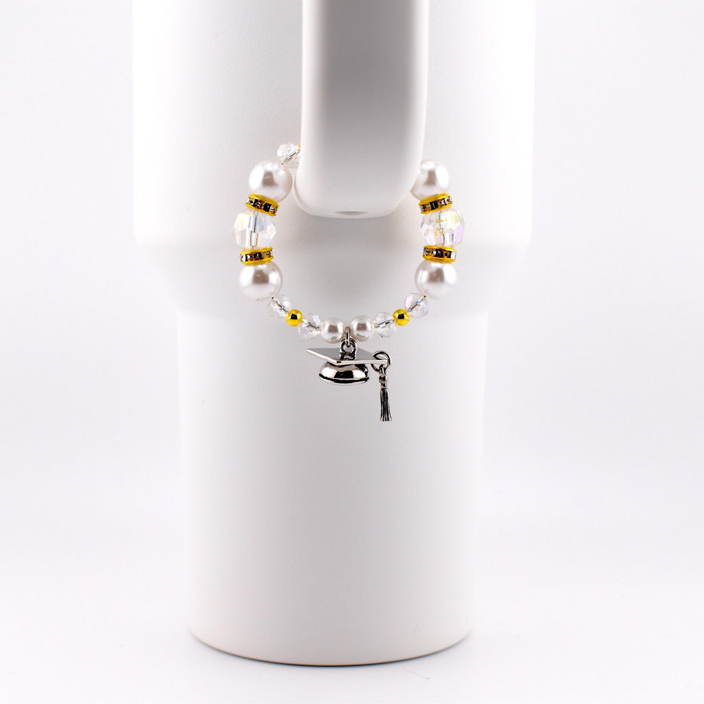 A white beaded cup charm with a graduation cap charm. The charm features white faux pearls, clear and gold-colored beads. The graduation cap charm is silver with a black tassel.