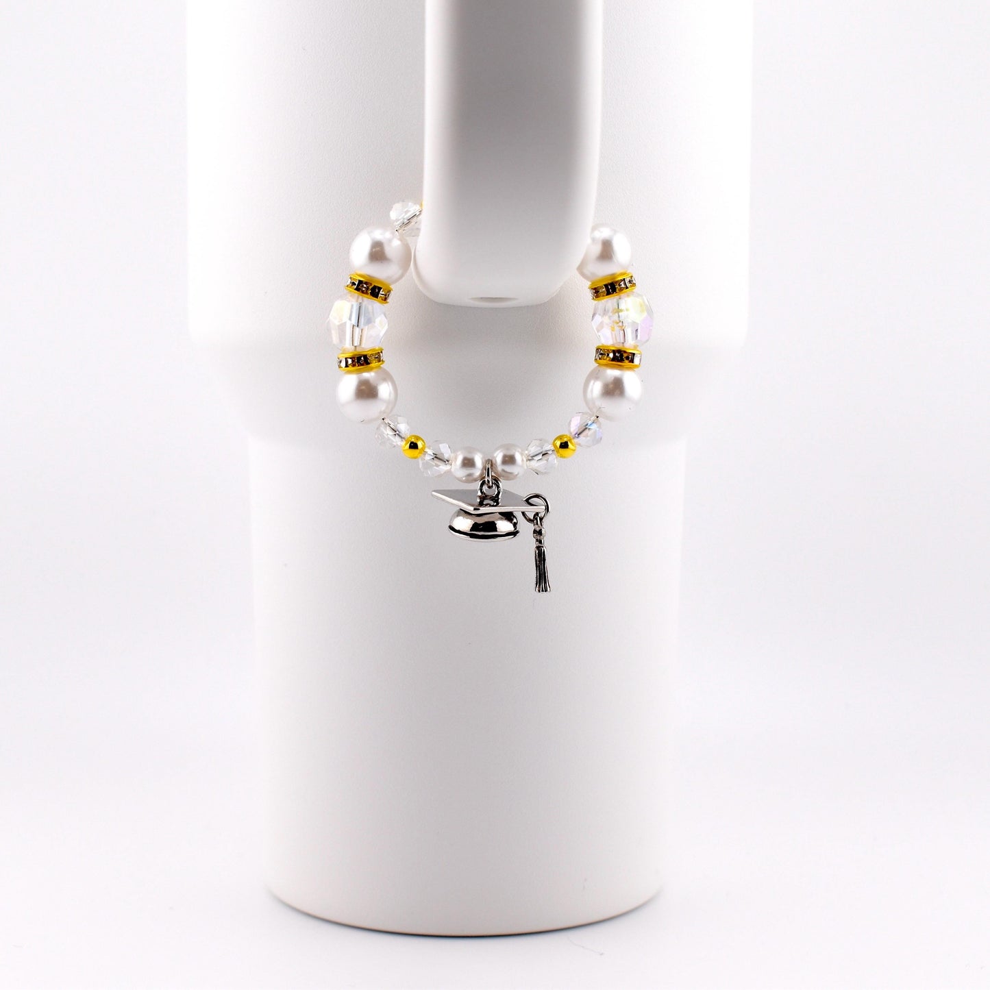 A white beaded cup charm with a graduation cap charm. The charm features white faux pearls, clear and gold-colored beads. The graduation cap charm is silver with a black tassel.