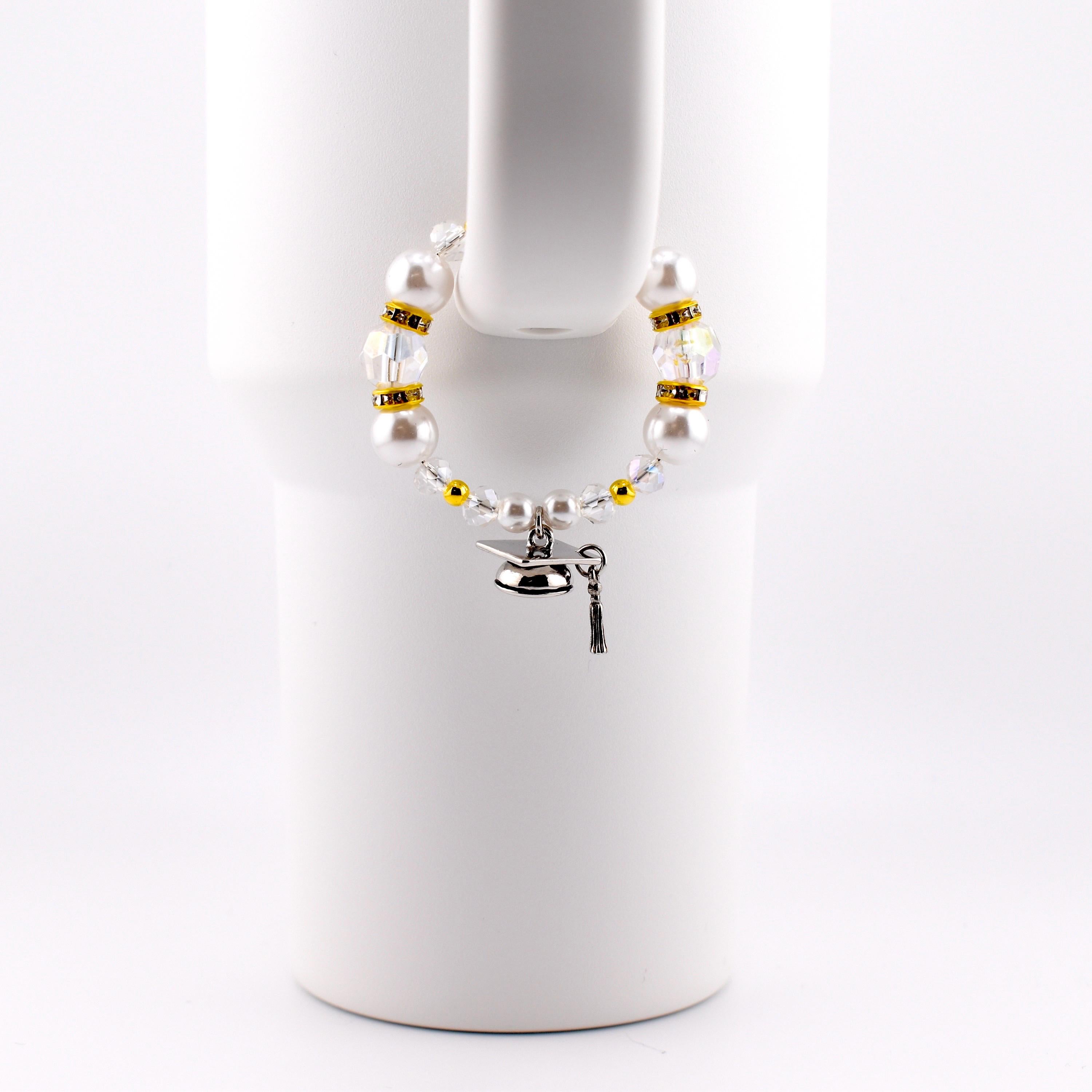 A white beaded cup charm with a graduation cap charm. The charm features white faux pearls, clear and gold-colored beads. The graduation cap charm is silver with a black tassel.