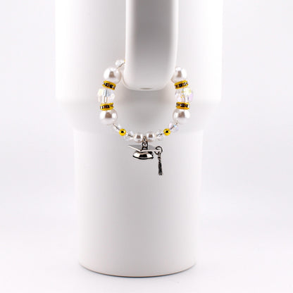 A white beaded cup charm with a graduation cap charm. The charm features white faux pearls, clear and gold-colored beads. The graduation cap charm is silver with a black tassel.