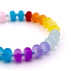 A rainbow-colored beaded bracelet with a variety of colors, including pink, blue, red, orange, yellow, and purple. The beads are round and smooth, strung together on a thin, clear cord. The bracelet is on a white background.