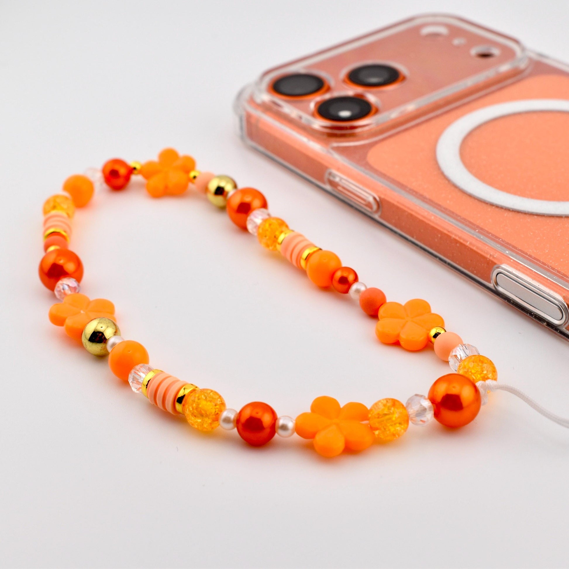 An orange beaded phone charm with flower and round bead accents. The charm is next to a clear phone case with an orange backing. The beads are in shades of orange, gold, and clear.
