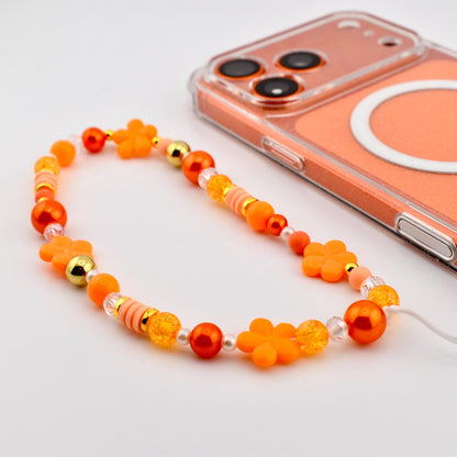 An orange beaded phone charm with flower and round bead accents. The charm is next to a clear phone case with an orange backing. The beads are in shades of orange, gold, and clear.