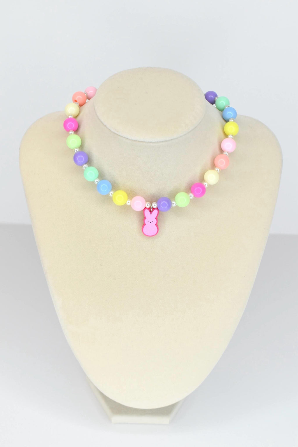 A colorful beaded necklace with pastel-colored beads in pink, yellow, green, blue, and purple. A pink bunny charm hangs from the center. The necklace is displayed on a mannequin.