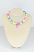 A colorful beaded necklace with pastel-colored beads in pink, yellow, green, blue, and purple. A pink bunny charm hangs from the center. The necklace is displayed on a mannequin.