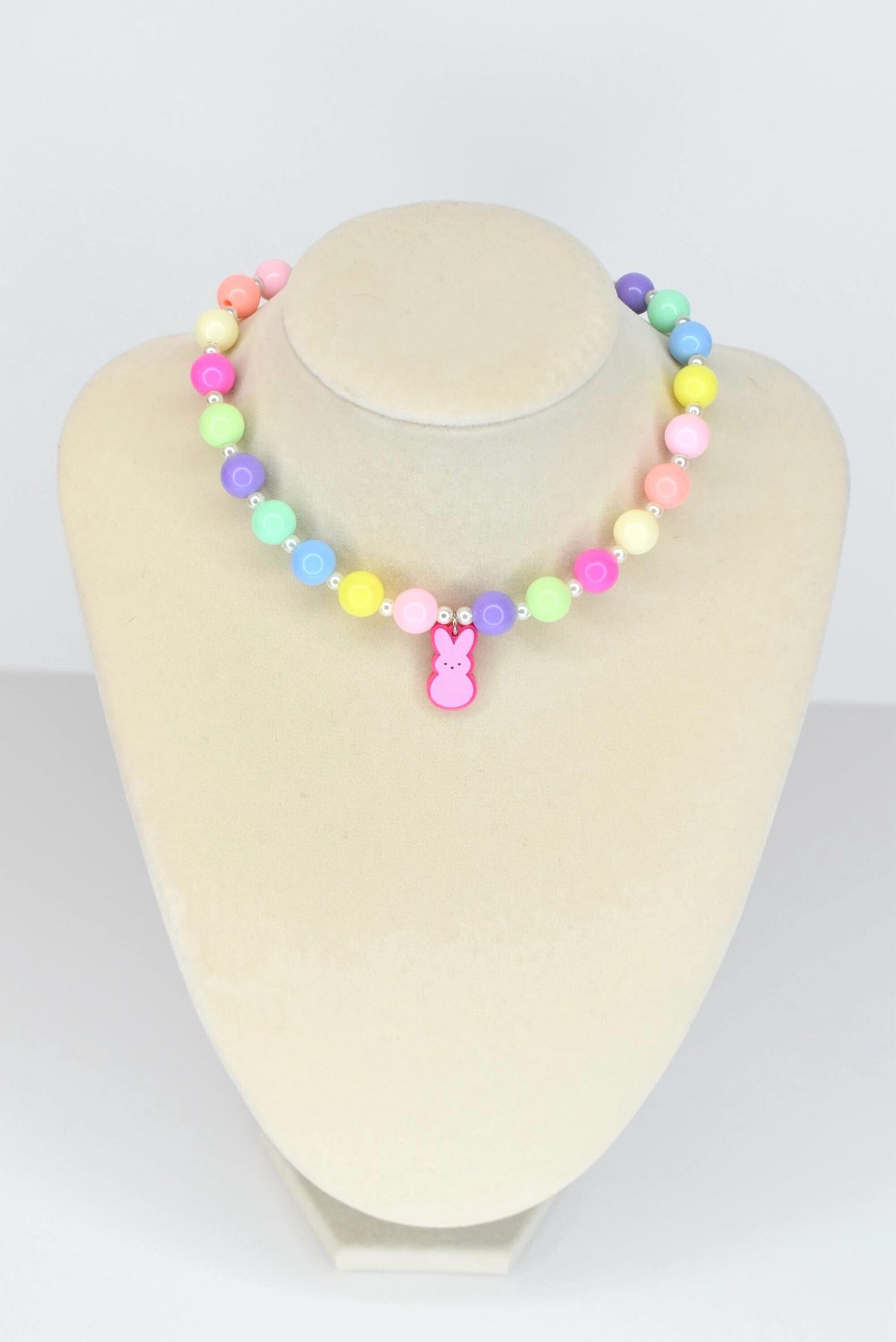 A colorful beaded necklace with pastel-colored beads in pink, yellow, green, blue, and purple. A pink bunny charm hangs from the center. The necklace is displayed on a mannequin.