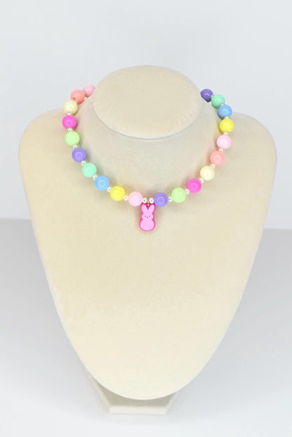 A colorful beaded necklace with pastel-colored beads in pink, yellow, green, blue, and purple. A pink bunny charm hangs from the center. The necklace is displayed on a mannequin.
