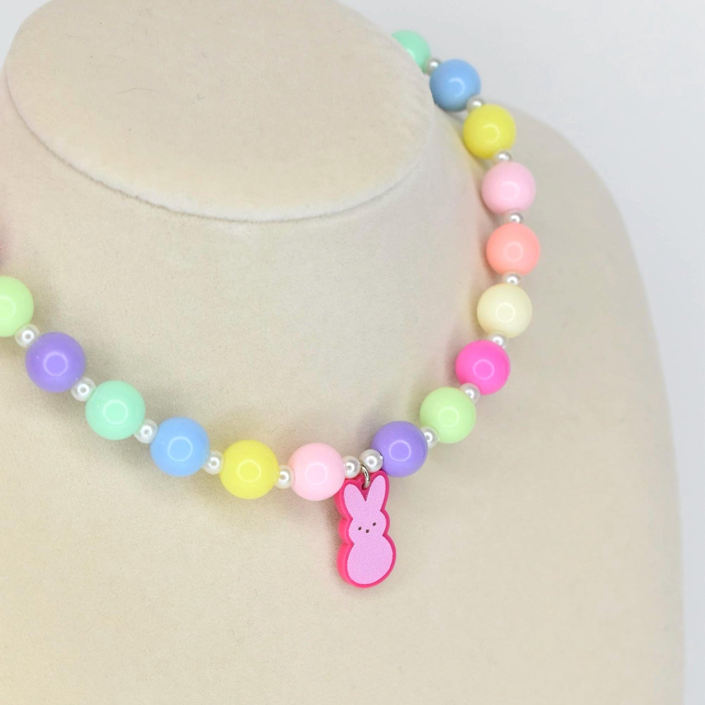 A colorful beaded necklace with pastel-colored beads in shades of pink, yellow, green, blue, and purple. A pink bunny charm hangs from the center. The necklace is displayed on a mannequin.