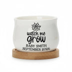 A white ceramic planter with a rounded shape, resting on a round wooden base. The planter features the text "watch me grow" in a script font, along with a succulent graphic and the name "BABY SMITH SEPTEMBER 2026".