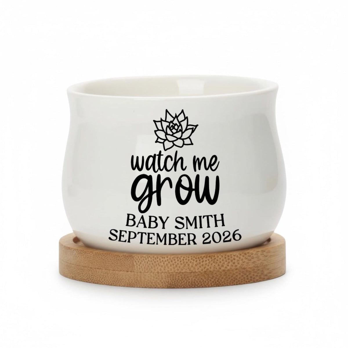 A white ceramic planter with a rounded shape, resting on a round wooden base. The planter features the text "watch me grow" in a script font, along with a succulent graphic and the name "BABY SMITH SEPTEMBER 2026".