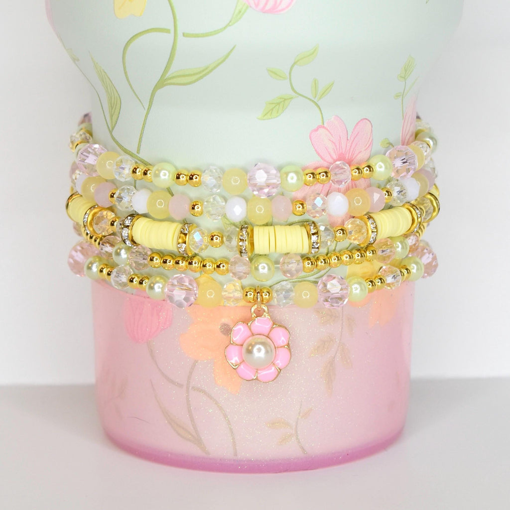 A stack of beaded bracelets in shades of yellow, pink, and clear, adorned with gold accents. The bracelets are wrapped around a decorative container with a floral design in pink and green. A flower charm with a pearl center hangs from the bracelets.