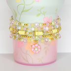 A stack of beaded bracelets in shades of yellow, pink, and clear, adorned with gold accents. The bracelets are wrapped around a decorative container with a floral design in pink and green. A flower charm with a pearl center hangs from the bracelets.