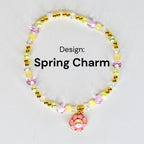 A beaded bracelet with the text "Spring Charm." The bracelet features a variety of beads in yellow, pink, clear, and gold colors. A pink flower charm with a pearl center hangs from the bracelet.
