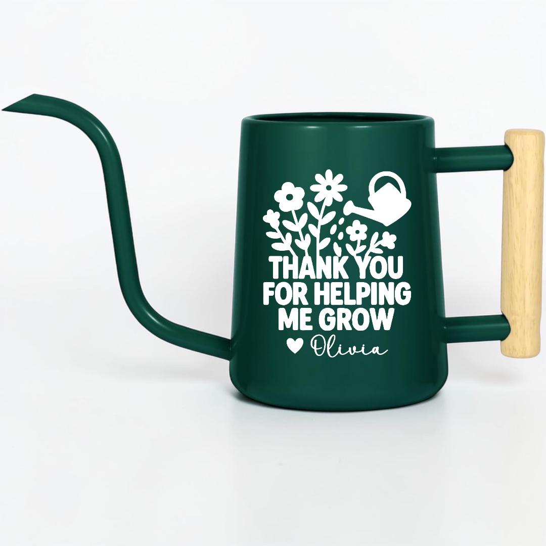 A dark green metal watering can with a long, curved spout and a wooden handle. The can features a white floral design and the text "THANK YOU FOR HELPING ME GROW" with the name "Olivia".