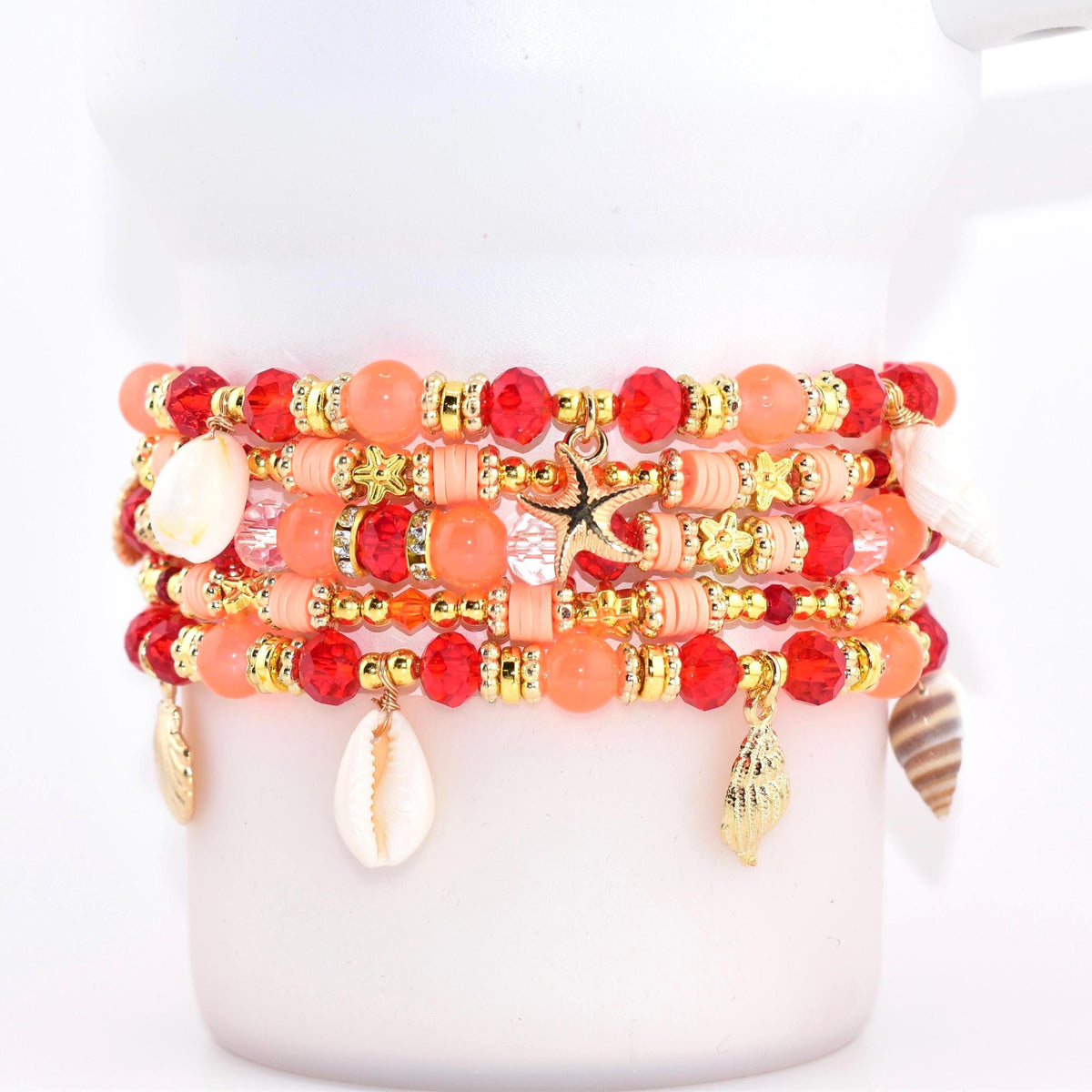 A stack of beaded bracelets in shades of red, coral, and gold. Each bracelet features a mix of beads, including faceted and round styles, with gold accents and seashell charms. A gold starfish charm hangs from one bracelet.