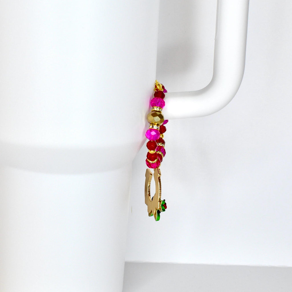 A beaded charm with a gold-colored horseshoe pendant and a small green and red accent. The charm features a mix of pink, red, and gold beads, attached to a white mug handle.