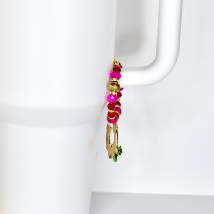 A beaded charm with a gold-colored horseshoe pendant and a small green and red accent. The charm features a mix of pink, red, and gold beads, attached to a white mug handle.
