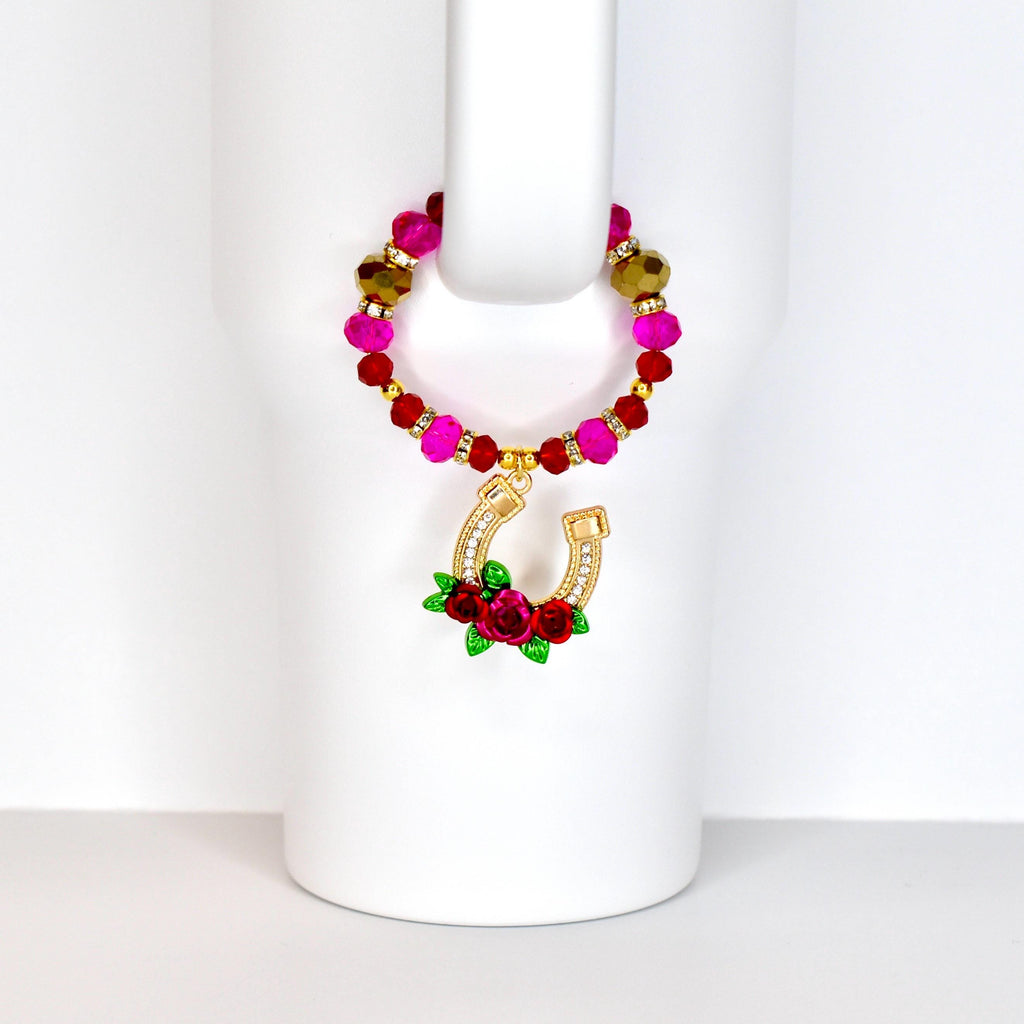 A beaded drink charm with a gold horseshoe pendant. The charm features a loop of pink, red, and gold beads. The horseshoe is adorned with clear crystals and a cluster of red roses with green leaves.