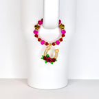 A beaded drink charm with a gold horseshoe pendant. The charm features a loop of pink, red, and gold beads. The horseshoe is adorned with clear crystals and a cluster of red roses with green leaves.