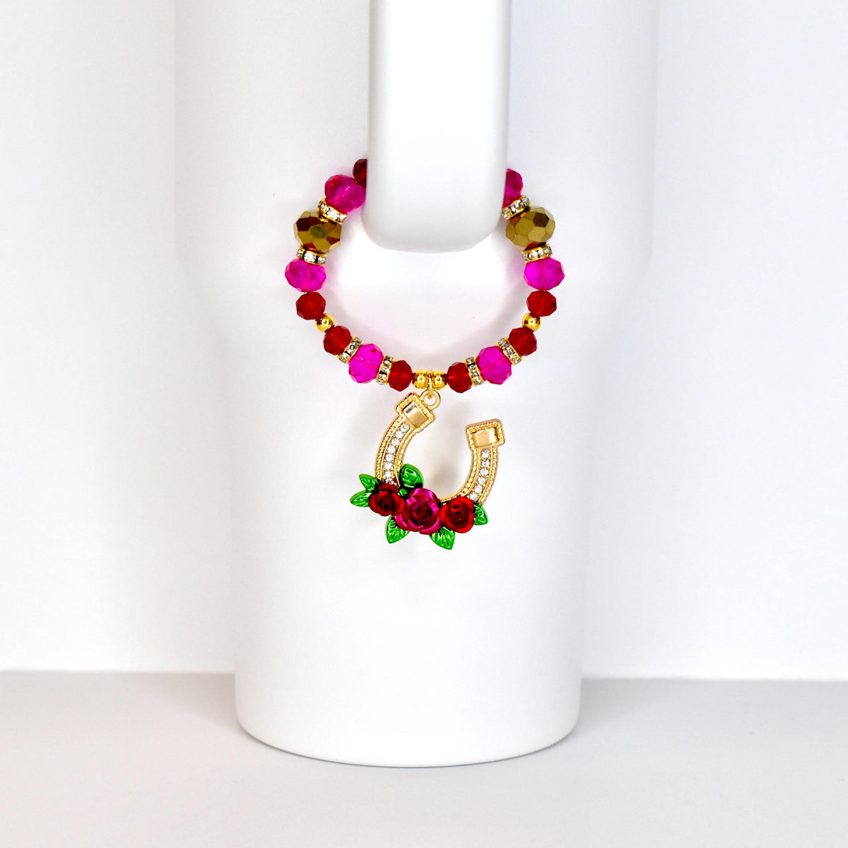 A beaded drink charm with a gold horseshoe pendant. The charm features a loop of pink, red, and gold beads. The horseshoe is adorned with clear crystals and a cluster of red roses with green leaves.