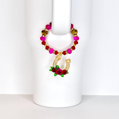 A beaded drink charm with a gold horseshoe pendant. The charm features a loop of pink, red, and gold beads. The horseshoe is adorned with clear crystals and a cluster of red roses with green leaves.