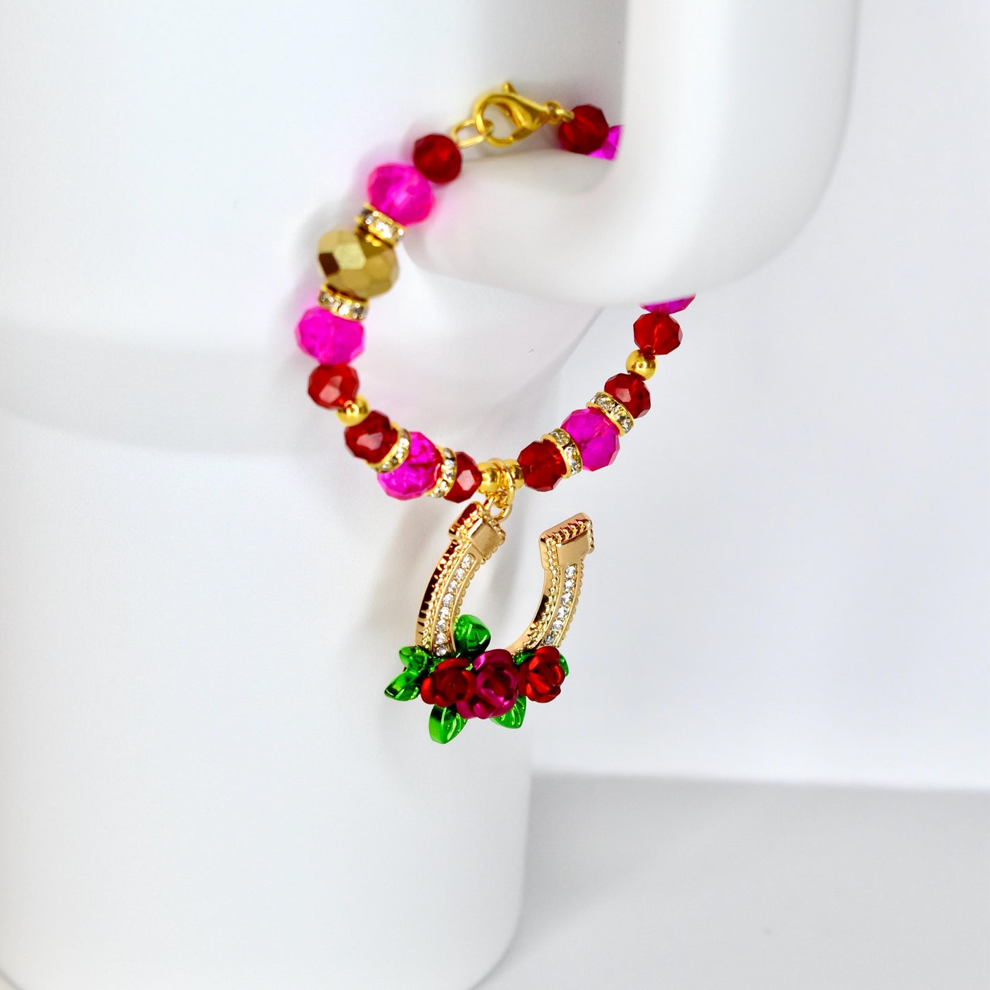 A beaded bracelet with a gold horseshoe charm. The bracelet features pink, red, and gold beads. The horseshoe charm is gold with clear crystals and red roses. The bracelet is displayed on a white surface.