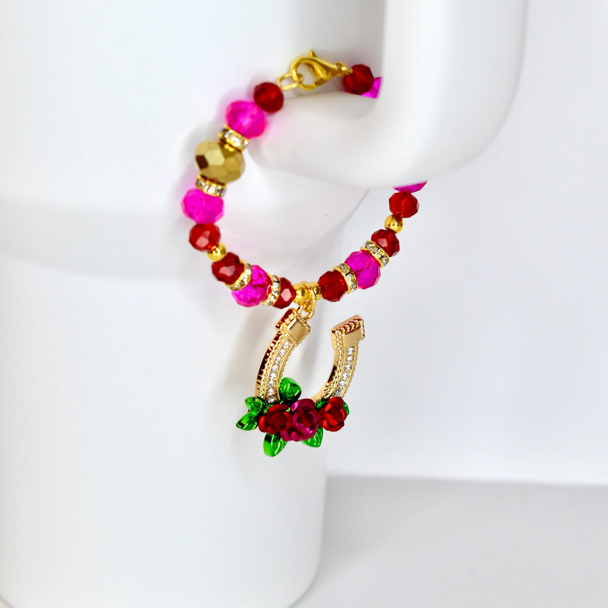 A beaded bracelet with a gold horseshoe charm. The bracelet features pink, red, and gold beads. The horseshoe charm is gold with clear crystals and red roses. The bracelet is displayed on a white surface.