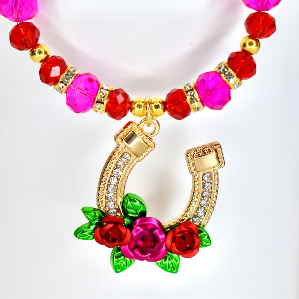 A tumbler handle charm featuring a gold-colored horseshoe pendant adorned with clear crystals and a cluster of red and pink roses with green leaves. The charm has a beaded strand of pink and red beads.