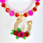 A tumbler handle charm featuring a gold-colored horseshoe pendant adorned with clear crystals and a cluster of red and pink roses with green leaves. The charm has a beaded strand of pink and red beads.