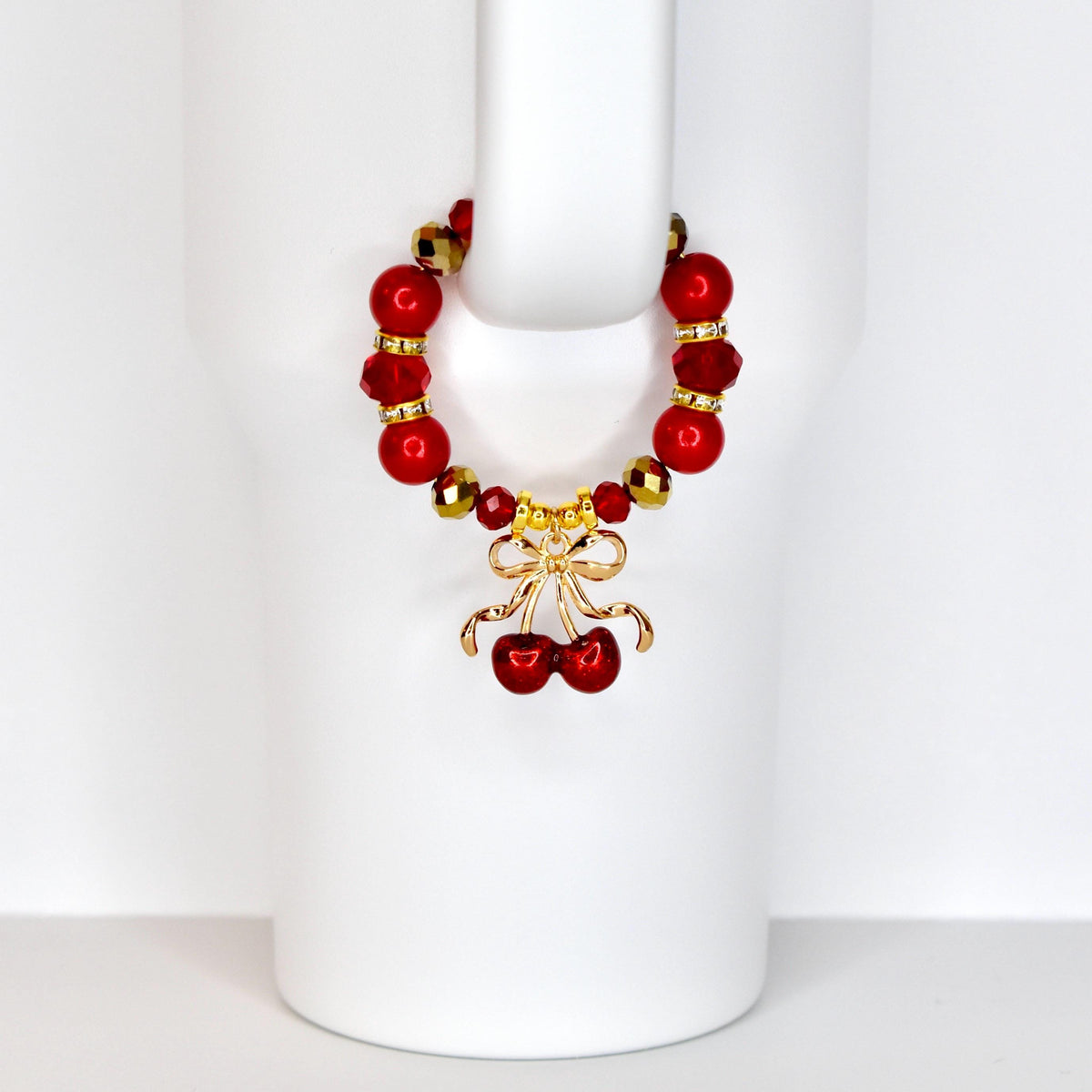 A red beaded tumbler handle bracelet with gold accents and a gold cherry charm. The bracelet features large red beads and smaller gold beads. The cherry charm has a bow and two cherries.