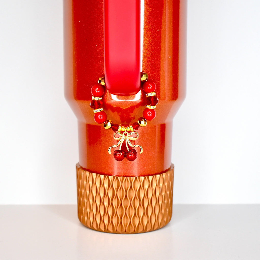 A red tumbler with a gold cherry charm and a decorative gold base. The charm features red beads and a gold bow with two red cherries. The tumbler has a red handle and a textured gold base.
