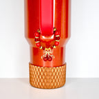 A red tumbler with a gold cherry charm and a decorative gold base. The charm features red beads and a gold bow with two red cherries. The tumbler has a red handle and a textured gold base.