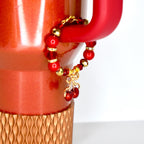A red beaded charm bracelet with gold accents and a cherry charm is attached to a red mug. The bracelet features red beads and gold-colored beads. The cherry charm is a gold bow with two red cherries.