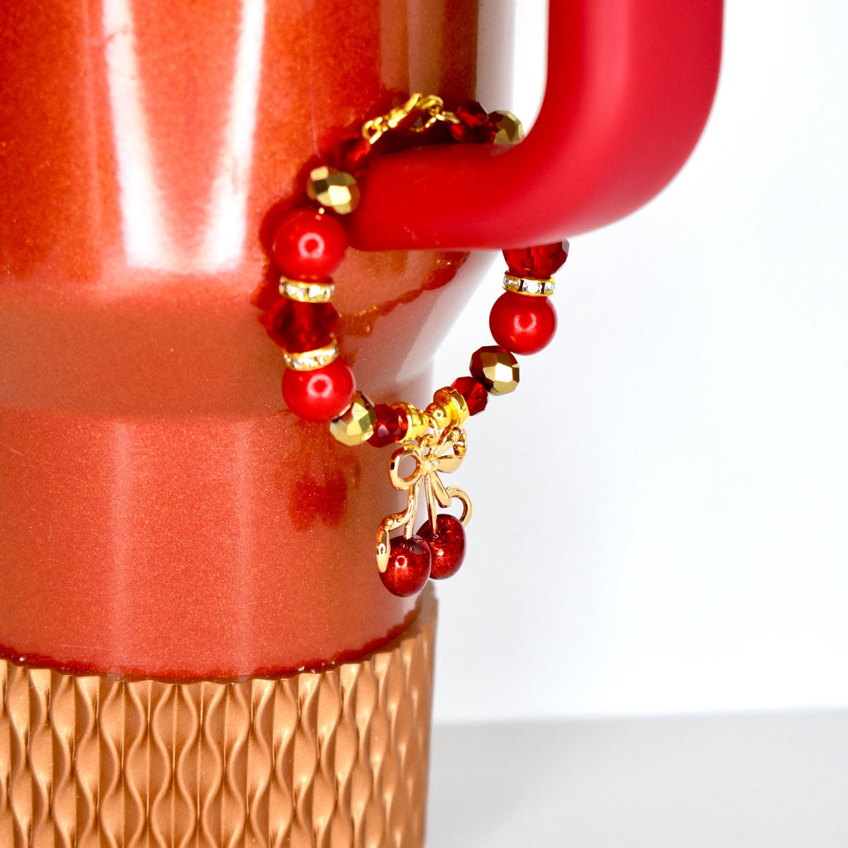 A red beaded charm bracelet with gold accents and a cherry charm is attached to a red mug. The bracelet features red beads and gold-colored beads. The cherry charm is a gold bow with two red cherries.