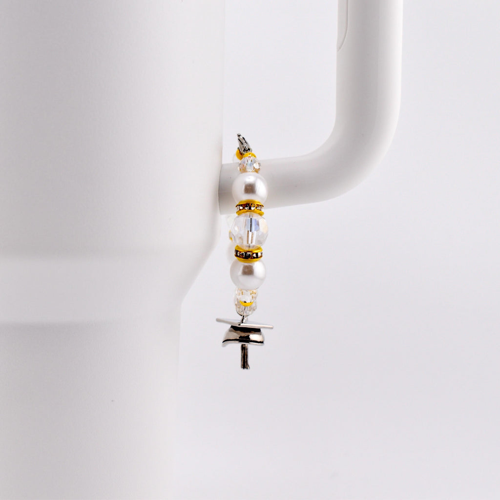 A decorative charm for a tumbler, featuring white pearls, clear beads, and gold accents. The charm is attached to a silver-colored metal piece with a T-shape. The charm is hanging on the handle of a white mug.