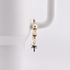 A decorative charm for a tumbler, featuring white pearls, clear beads, and gold accents. The charm is attached to a silver-colored metal piece with a T-shape. The charm is hanging on the handle of a white mug.