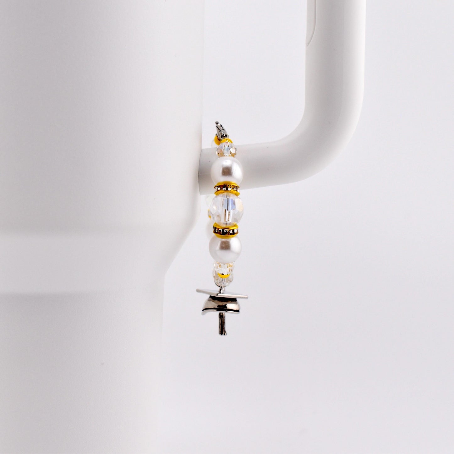 A decorative charm for a tumbler, featuring white pearls, clear beads, and gold accents. The charm is attached to a silver-colored metal piece with a T-shape. The charm is hanging on the handle of a white mug.