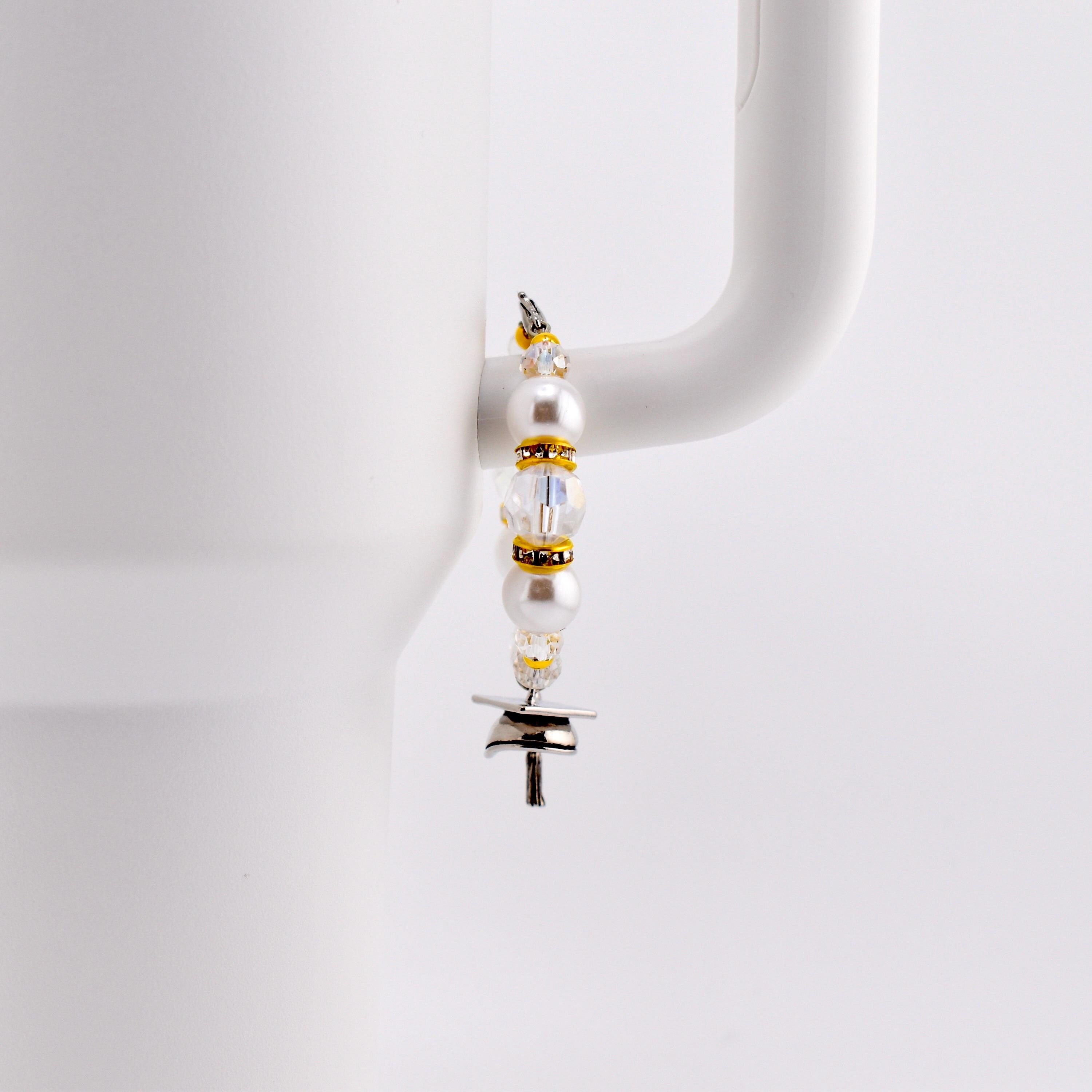 A decorative charm for a tumbler, featuring white pearls, clear beads, and gold accents. The charm is attached to a silver-colored metal piece with a T-shape. The charm is hanging on the handle of a white mug.