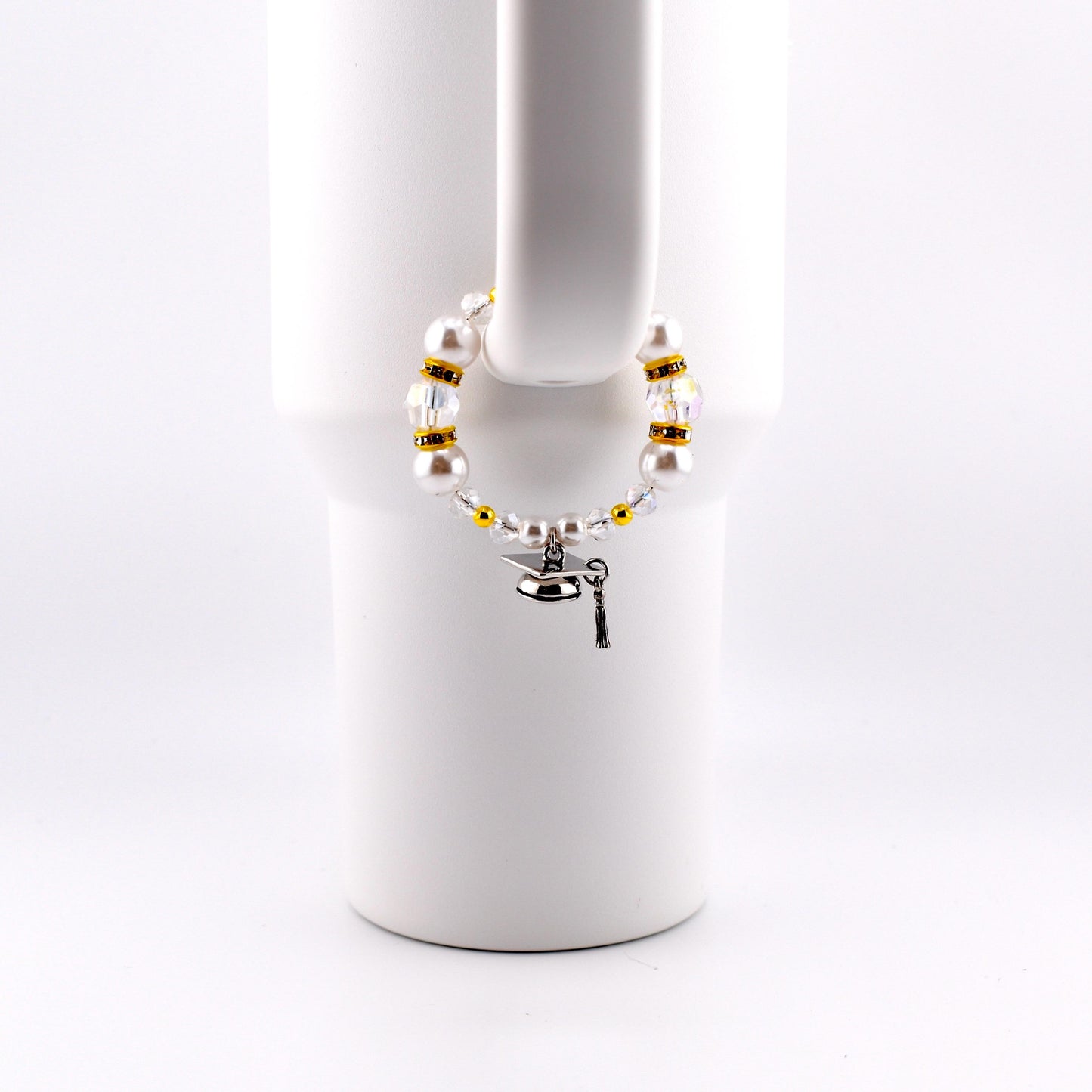 A graduation-themed bracelet with white faux pearls, gold beads, and clear crystals. A small silver graduation cap charm and key charm dangle from the bracelet. The bracelet is displayed on a white mug handle.