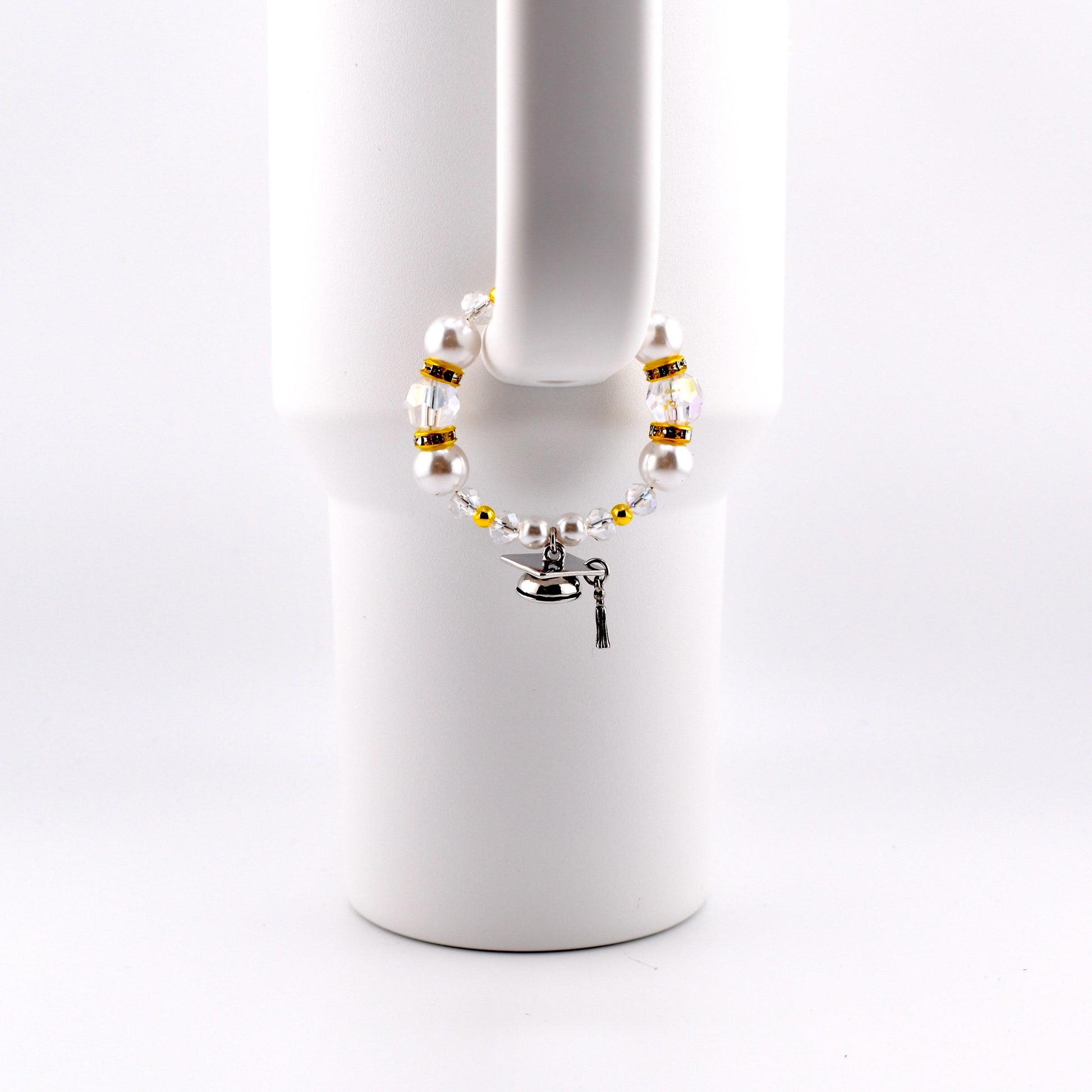 A graduation-themed bracelet with white faux pearls, gold beads, and clear crystals. A small silver graduation cap charm and key charm dangle from the bracelet. The bracelet is displayed on a white mug handle.