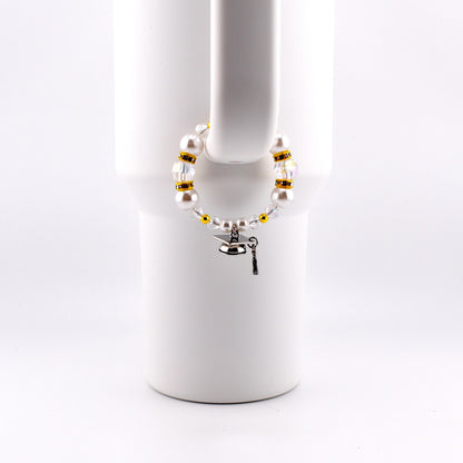 A graduation-themed bracelet with white faux pearls, gold beads, and clear crystals. A small silver graduation cap charm and key charm dangle from the bracelet. The bracelet is displayed on a white mug handle.