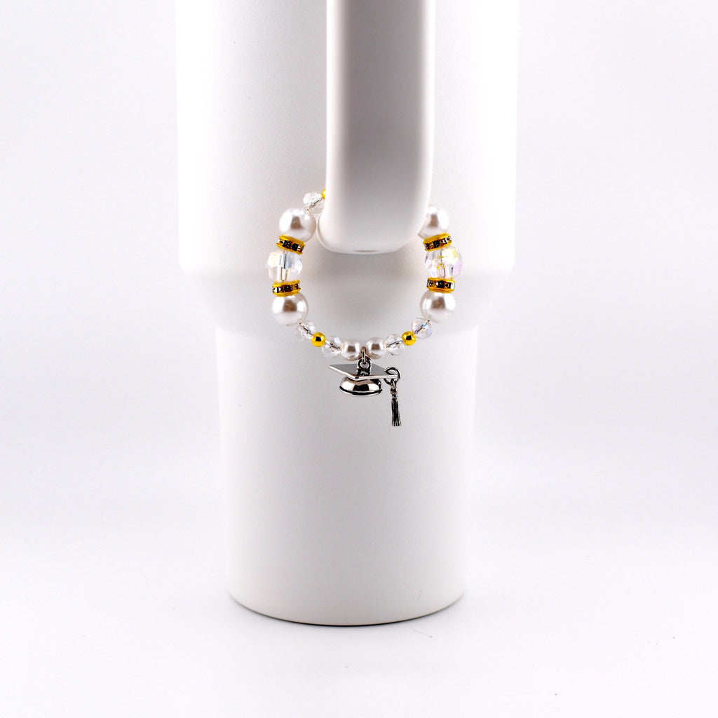 A graduation-themed bracelet with white faux pearls, gold beads, and clear crystals. A small silver graduation cap charm and key charm dangle from the bracelet. The bracelet is displayed on a white mug handle.