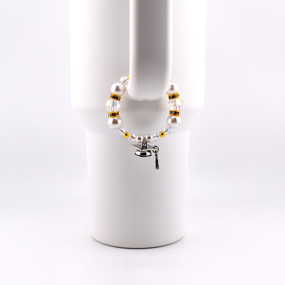 A graduation-themed bracelet with white faux pearls, gold beads, and clear crystals. A small silver graduation cap charm and key charm dangle from the bracelet. The bracelet is displayed on a white mug handle.