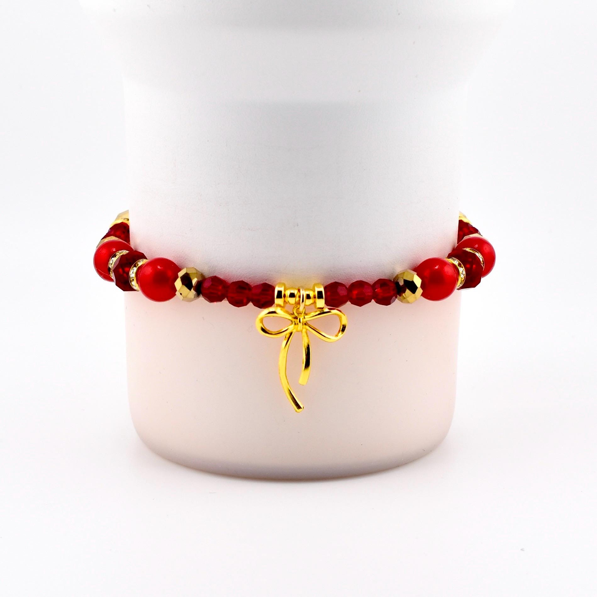 A red beaded bracelet with gold accents and a gold bow charm. The bracelet features large red beads, smaller red beads, and gold-colored spacers. The bow charm hangs from the center of the bracelet.