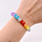 A rainbow-colored beaded bracelet with heart-shaped red beads. The bracelet features beads in shades of purple, blue, white, red, orange, yellow, and green. The bracelet is worn on a wrist.