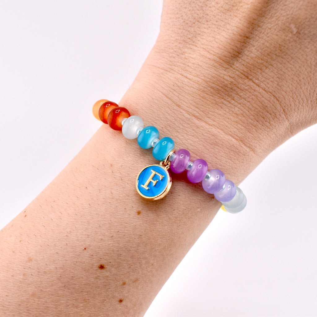A beaded bracelet with a rainbow color scheme. The bracelet features a gold charm with a blue enamel background and the letter "F". The beads are various colors, including red, orange, white, blue, purple, and yellow.