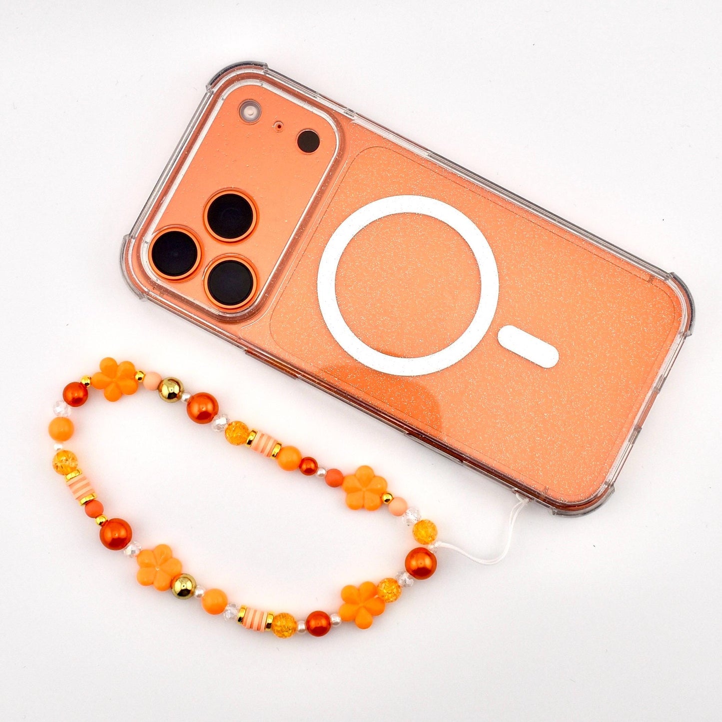 An orange phone with a clear border and a white circular design. A beaded phone strap in shades of orange, yellow, and gold is attached. The strap features flower and bead accents.