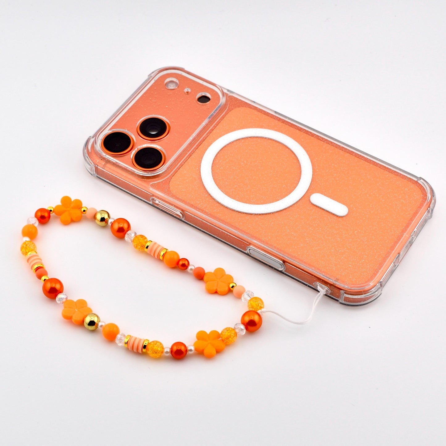 An orange phone with a clear border and a white circular design. A beaded phone strap in shades of orange, yellow, and gold features flower and bead accents. The phone and strap are on a white surface.
