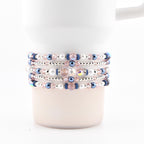 A white tumbler with several beaded bracelets wrapped around it. The bracelets feature a mix of pink, blue, and white beads, with silver accents. The beads are arranged in a repeating pattern, creating a decorative accessory.
