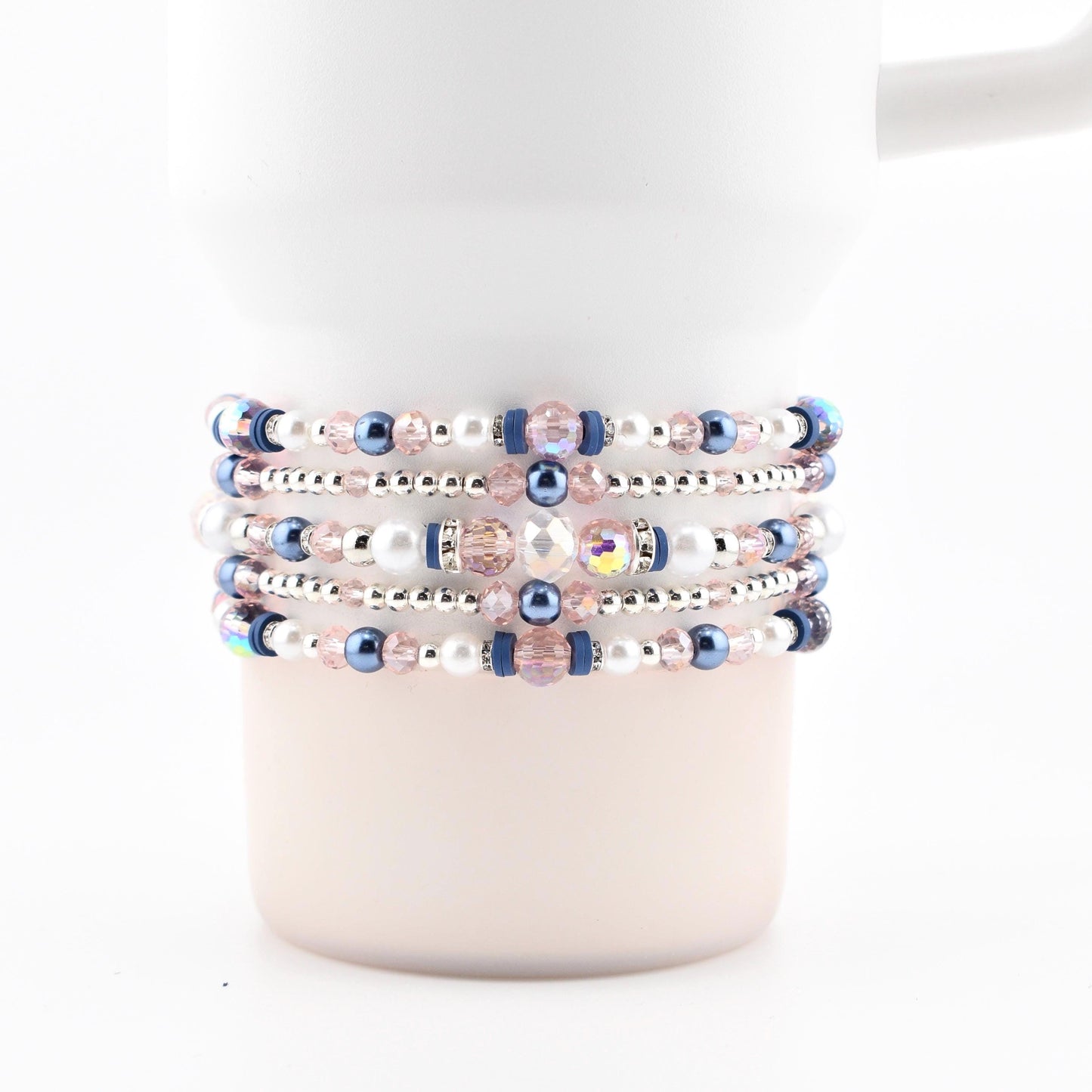 A white tumbler with several beaded bracelets wrapped around it. The bracelets feature a mix of pink, blue, and white beads, with silver accents. The beads are arranged in a repeating pattern, creating a decorative accessory.